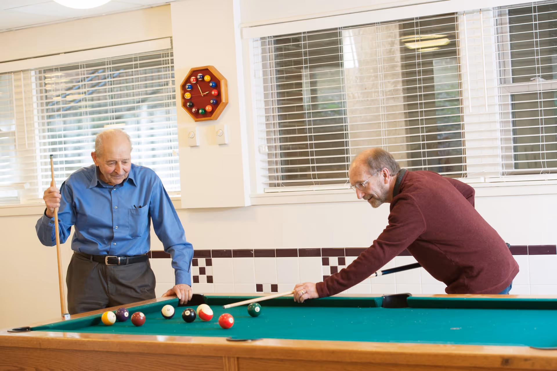 Two elderly men playing pool in a well-lit room with large windows and white blinds. One man in a blue shirt is standing and holding a pool cue, while the other man in a maroon sweater is leaning over the pool table aiming a shot. A wall clock designed like a billiard rack with pool balls is mounted on the wall behind them.