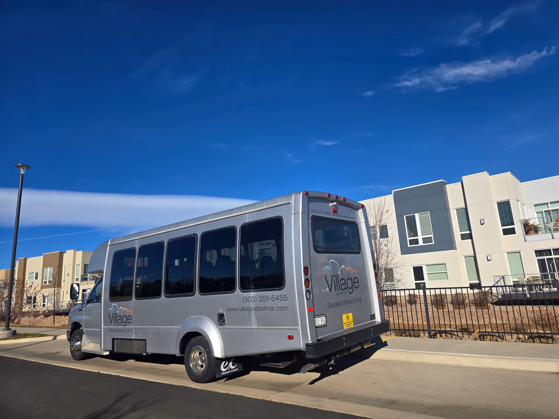 A silver Village at Belmar shuttle bus parked on the street in front of a modern senior living building under a blue sky.