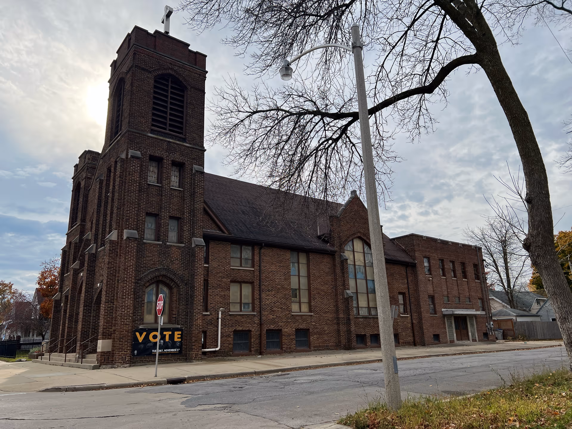 Exterior view of a large brick building with a tall tower and cross on top, likely a church, on a cloudy day. There is a stop sign and a sign that says 'VOTE' near the corner of the building. Leafless trees and a streetlight are visible in the foreground.