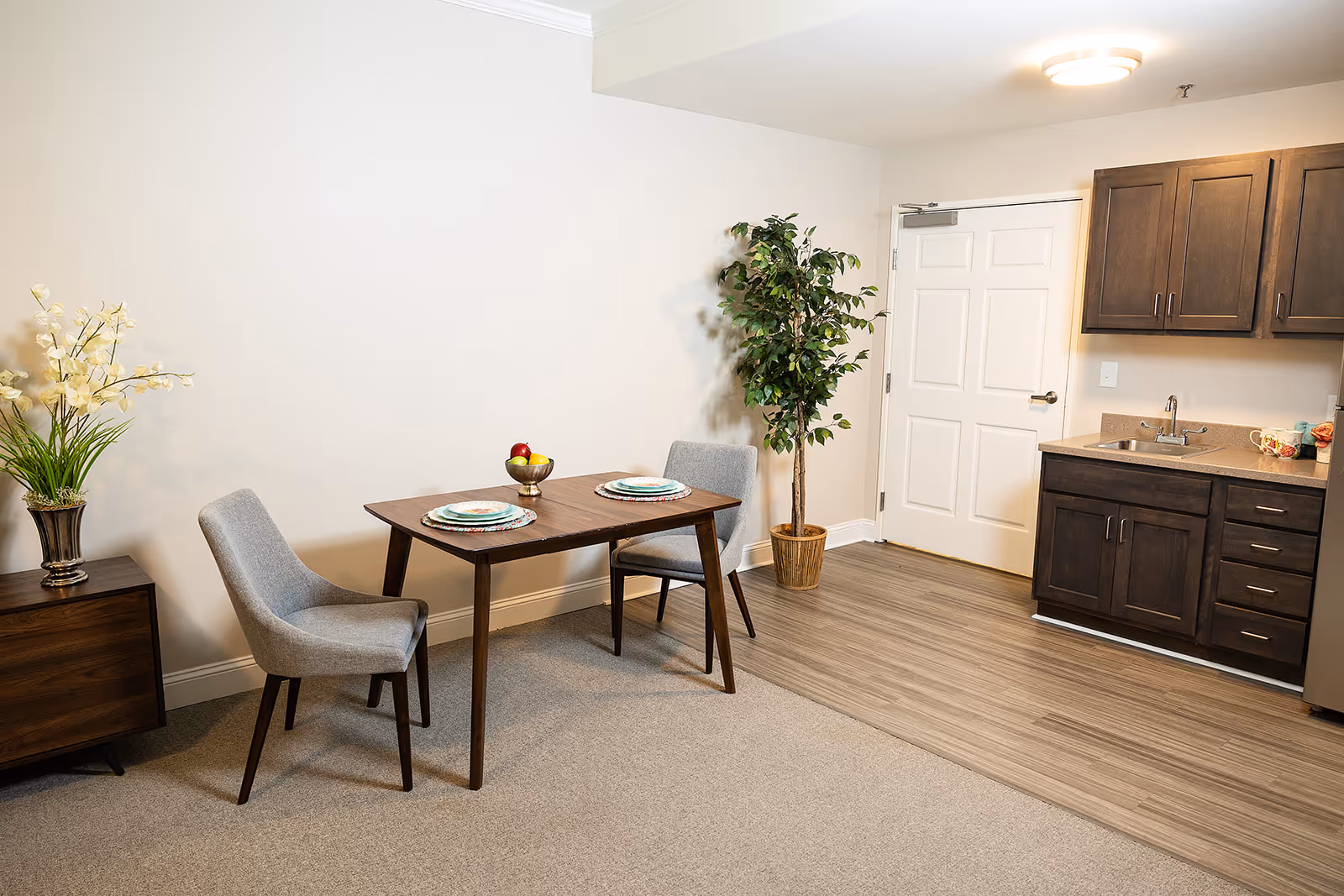 A small dining area with a wooden table set for two with plates and a bowl of fruit, two gray upholstered chairs, a potted plant in the corner, and a kitchenette with dark wood cabinets, a sink, and a countertop. The floor transitions from carpet to wood near the kitchenette, and there is a white door in the background.