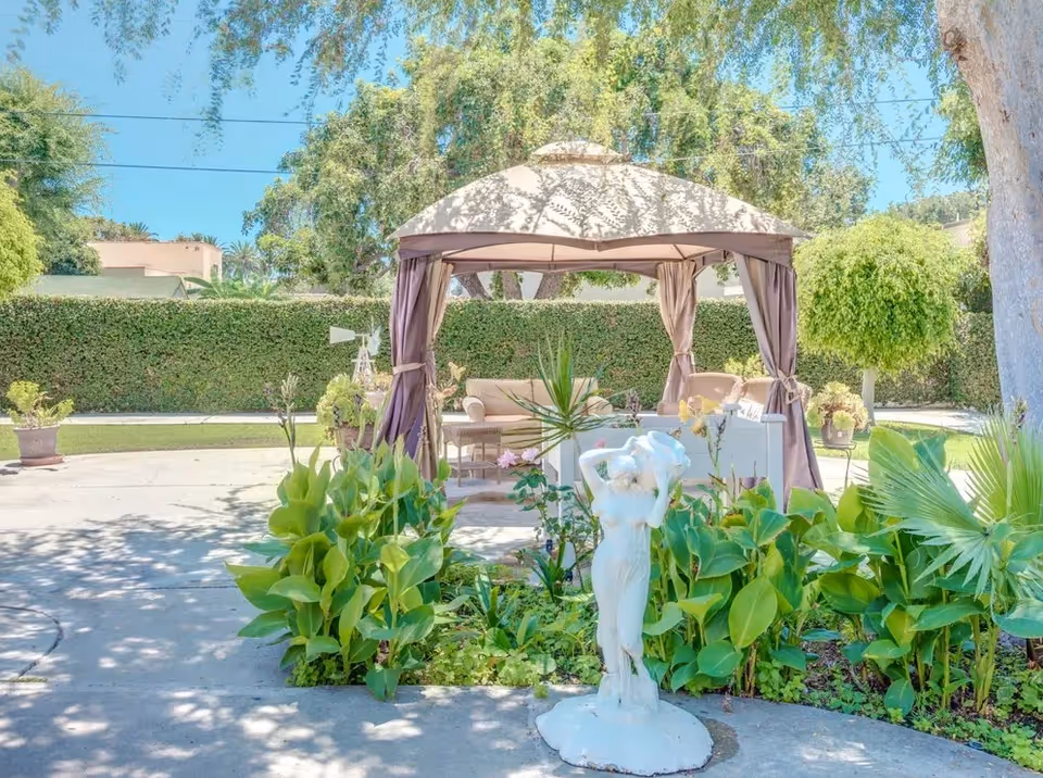 Outdoor garden area with a white statue of a woman in the foreground surrounded by green plants. Behind the statue is a beige gazebo with curtains tied back, containing outdoor seating including sofas and a table. Trees and a tall hedge form the background under a clear blue sky.