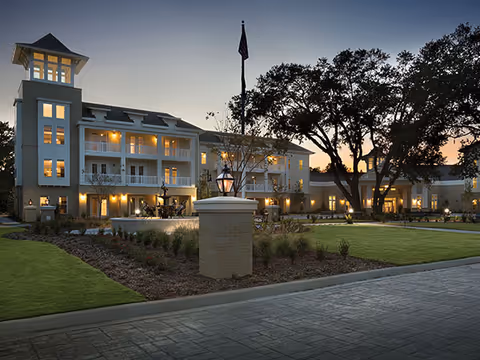 Exterior view of South Bay At Mount Pleasant senior living facility at dusk, showing a well-lit multi-story building with balconies, a flagpole, large trees, and landscaped grounds with a paved driveway in the foreground.