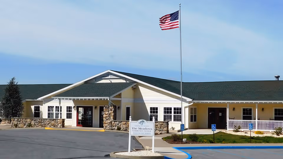 Exterior view of a single-story senior living facility building with a green roof and light yellow siding. There is a flagpole with an American flag in front of the building, a paved driveway, and a sign that reads 'The Meadows Community Center'. The sky is clear with some light clouds.