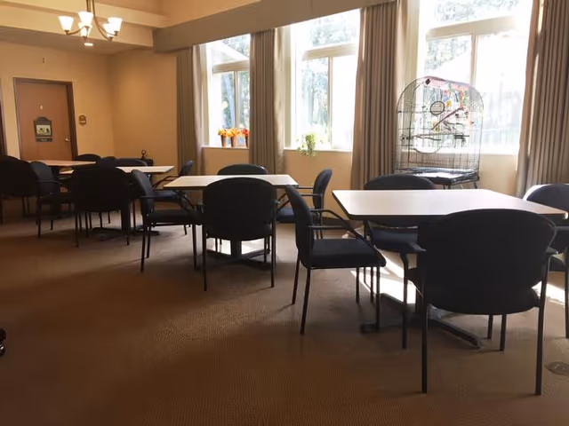 Sunlit communal dining/activity room with several tables and chairs by large windows and a birdcage.
