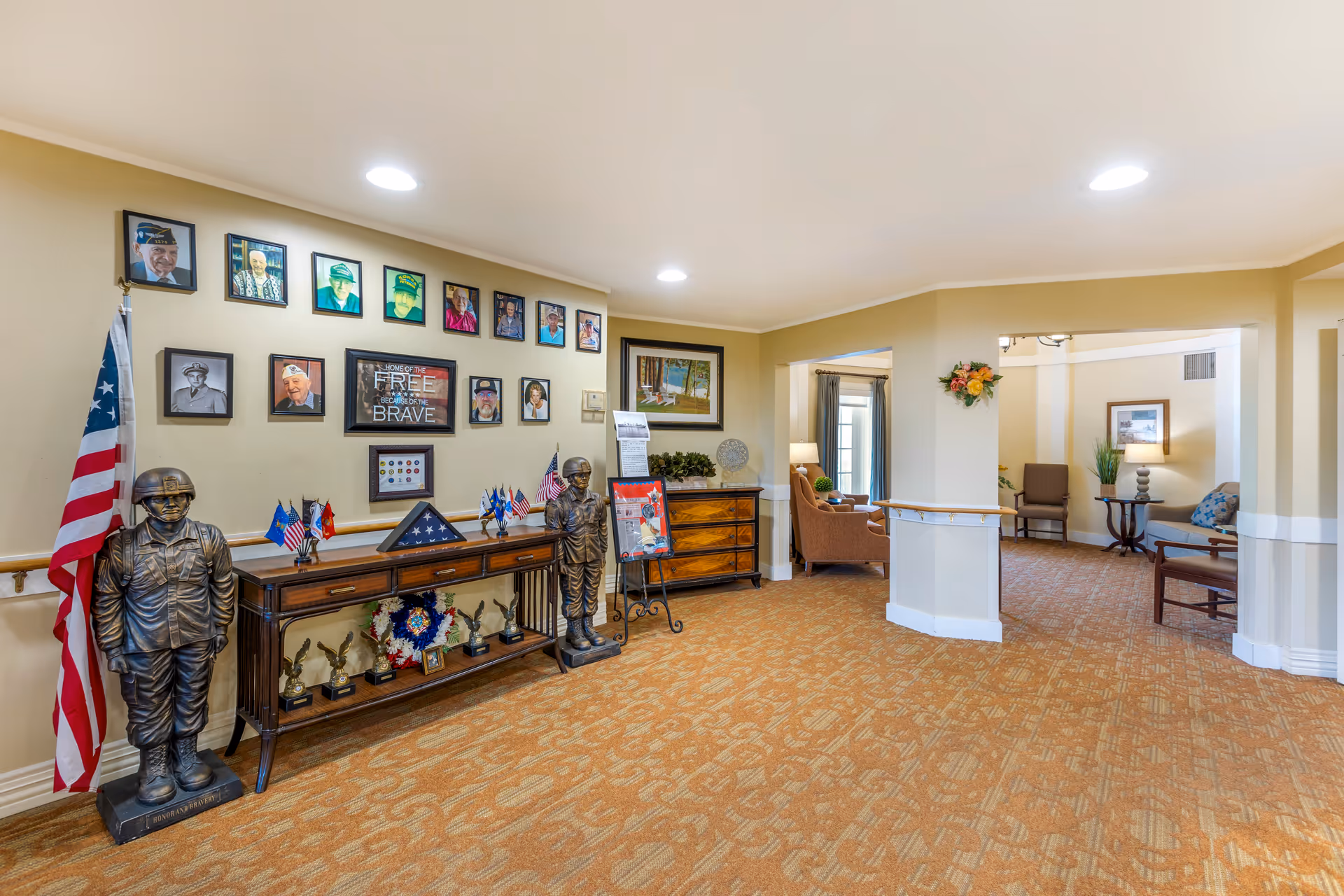 Interior view of a senior living facility hallway with a military tribute display on the left wall, featuring framed photos of veterans, two soldier statues, American flags, and various awards on a wooden table. The hallway leads to a sitting area with chairs, tables, lamps, and decorative plants.