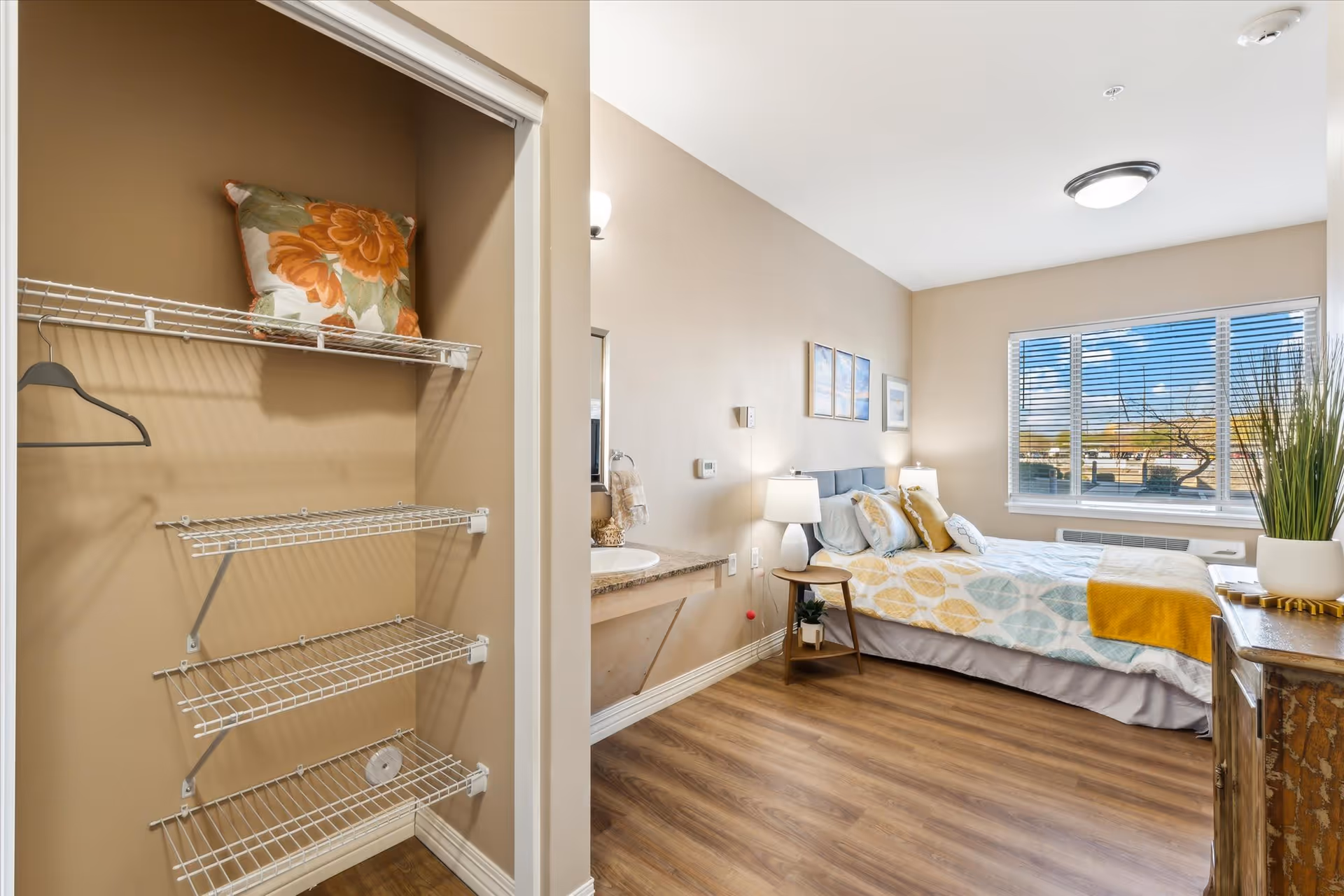 A bright bedroom with a large window showing an outdoor view. The room features a bed with patterned bedding and multiple pillows, two bedside tables with lamps, a wooden dresser with a potted plant, and a small open closet with wire shelves and a floral pillow on the top shelf. The walls are painted beige and the floor is wood.