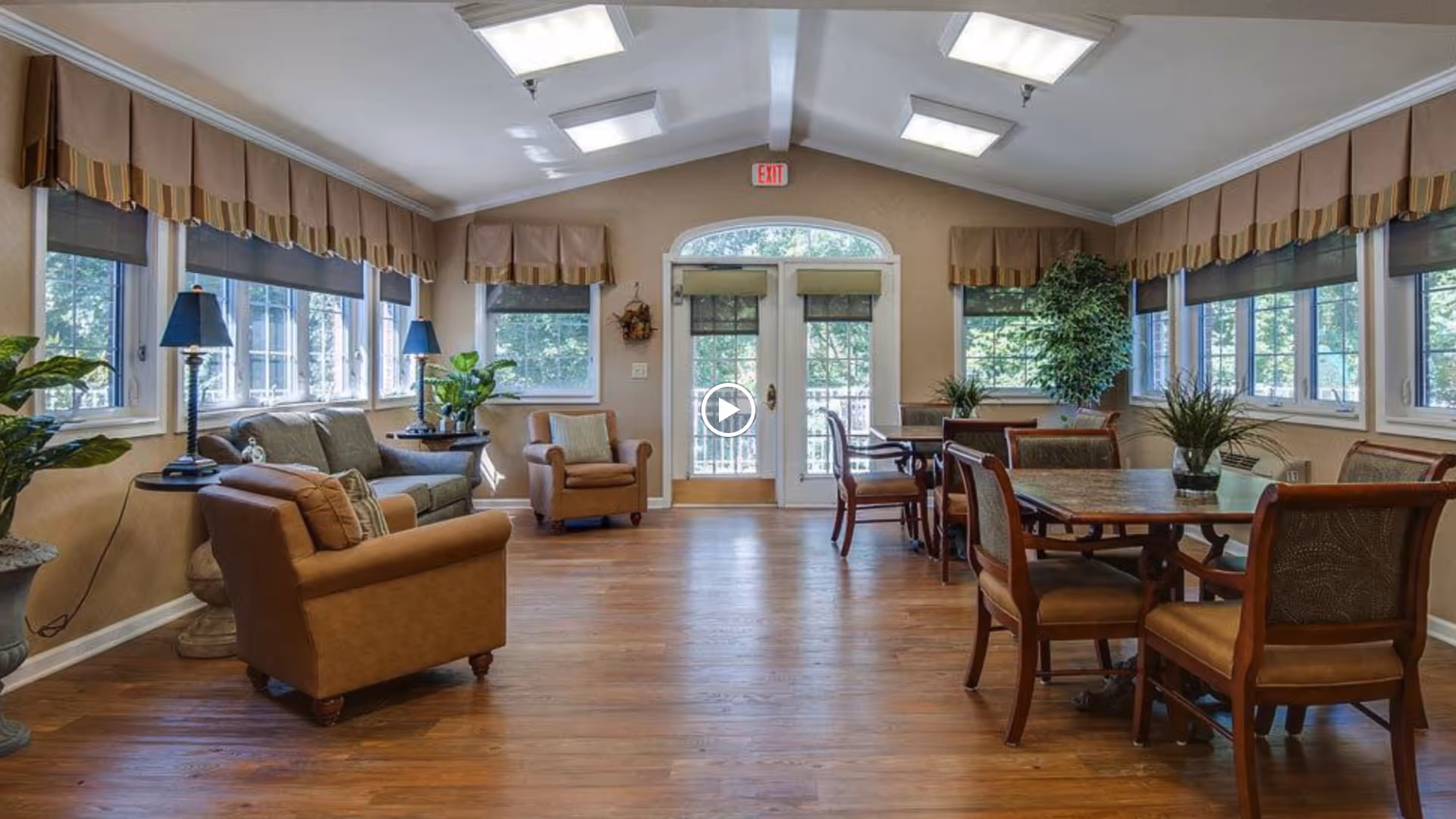 A bright, spacious common area with wooden flooring, multiple windows with valances, and skylights. The room is furnished with comfortable armchairs, a sofa, side tables with lamps, and dining tables with chairs. There are several potted plants adding greenery to the space, and double glass doors leading outside.