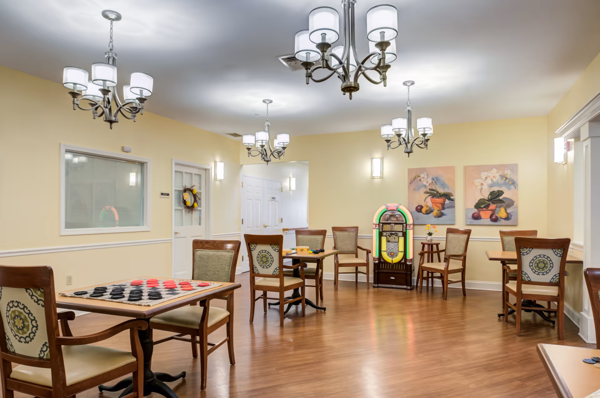 Bright communal activity/dining room with several tables and chairs, a jukebox, and wall paintings.