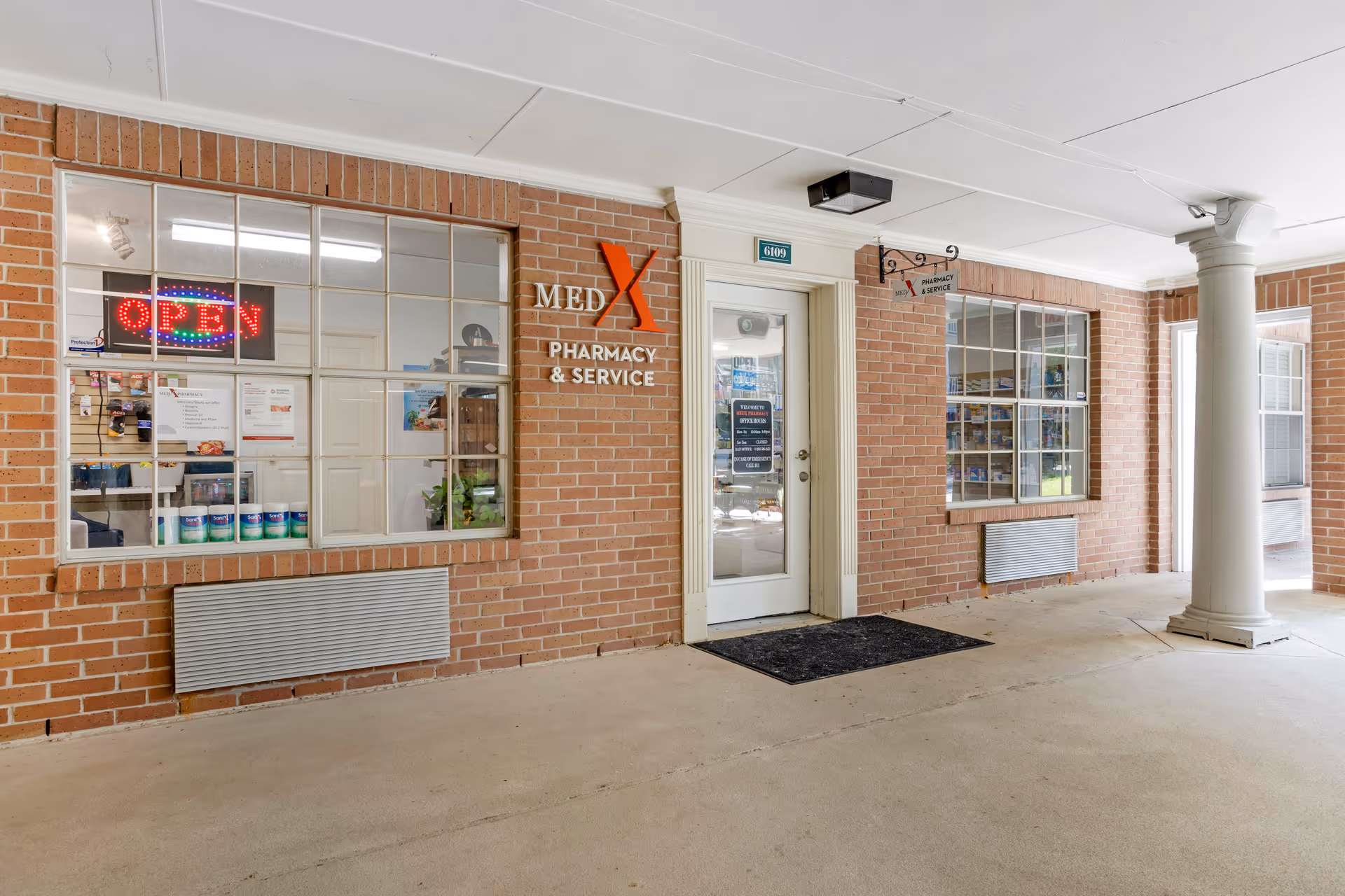 Entrance to MedX Pharmacy & Service located in a brick building with large windows displaying various products and an illuminated open sign. The door has a sign with hours and information, and there is a black mat in front of the door under a covered walkway with a white column.