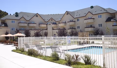 Outdoor swimming pool area with white fencing, lounge chairs, and umbrellas in front of a large multi-story residential building under a clear blue sky.