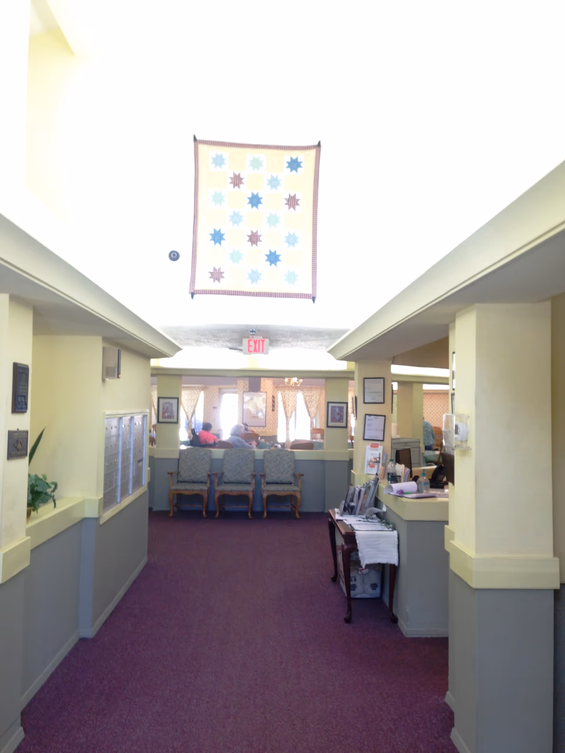 Interior hallway of an assisted living facility with purple carpet and pale yellow walls. There are three upholstered chairs against a half wall at the end of the hallway, with a quilt hanging above and an exit sign visible. On the right side, there is a reception desk with papers and a hand sanitizer dispenser. On the left side, there are mailboxes and a small plant.
