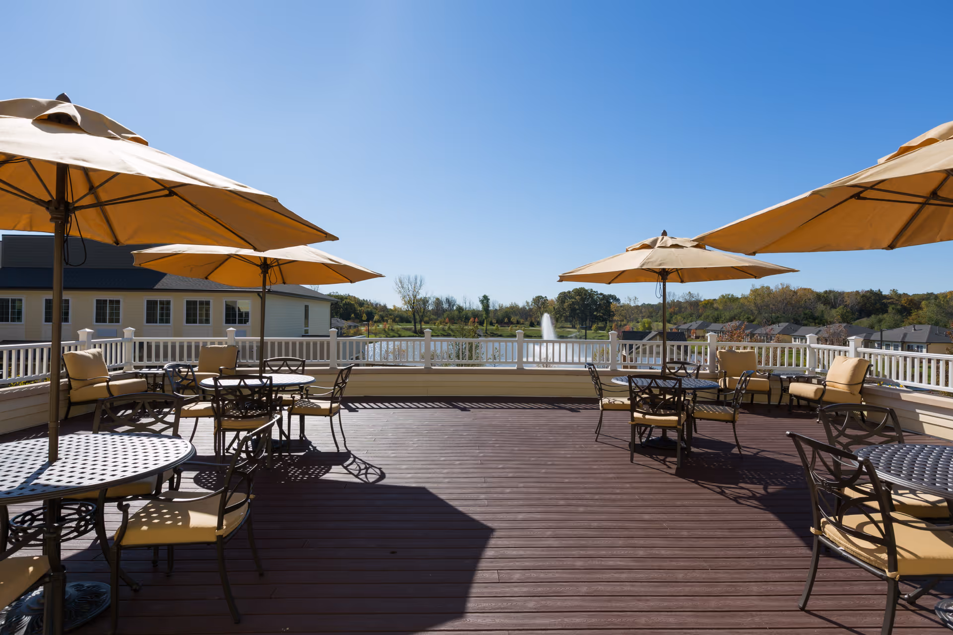 Outdoor patio area at Green Oaks Senior Living with several round metal tables and cushioned chairs under large beige umbrellas. The patio overlooks a scenic view of a pond with a water fountain and trees in the background under a clear blue sky.