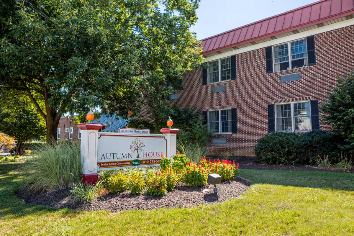 Exterior view of Autumn House East senior living community building with a brick facade, surrounded by green grass, bushes, and trees. A white sign with red and green accents displaying the facility name and contact information is prominently placed in front of the building.