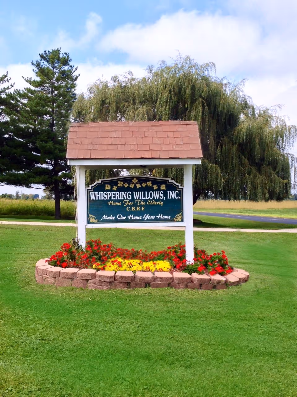 A wooden sign for Whispering Willows, Inc. stands on a grassy lawn with a flower bed of red and yellow flowers at its base. Behind the sign are tall trees and a partly cloudy sky.