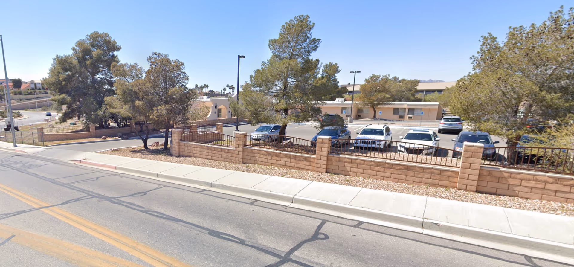 Exterior view of Mountain View Care Center showing a parking lot with several cars, a low brick wall with metal fencing, trees, and a clear blue sky.
