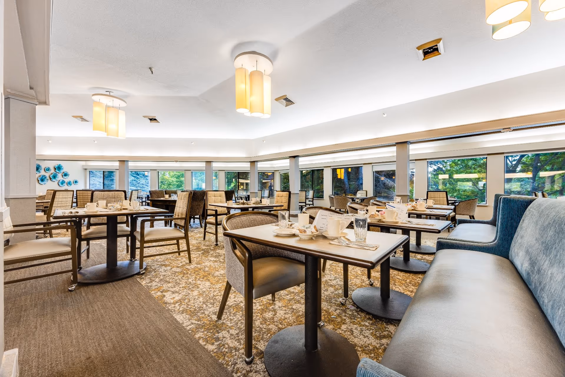 A bright and spacious dining room with multiple tables and chairs arranged neatly. Each table is set with cups, plates, glasses, and utensils. Large windows line the walls, allowing natural light to fill the room and offering views of greenery outside. The ceiling has modern light fixtures, and the floor is carpeted with a patterned design.