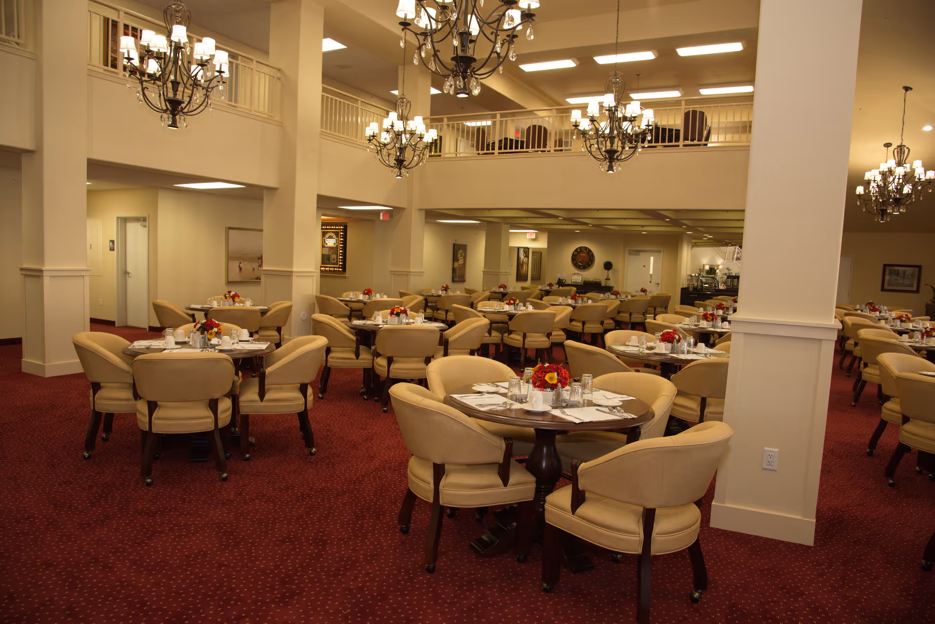 Spacious formal dining room with round tables set for meals, beige upholstered chairs, chandeliers, and red carpet.