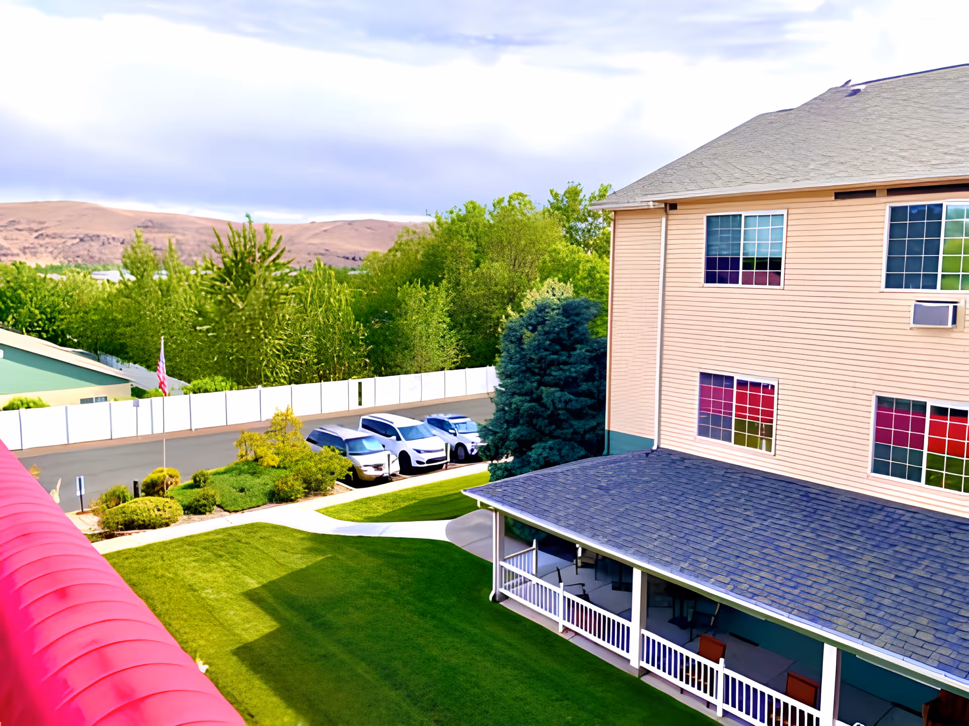 View of a senior living facility's outdoor area featuring a well-maintained green lawn, a covered porch with seating, several parked cars, and a backdrop of trees and hills under a partly cloudy sky.