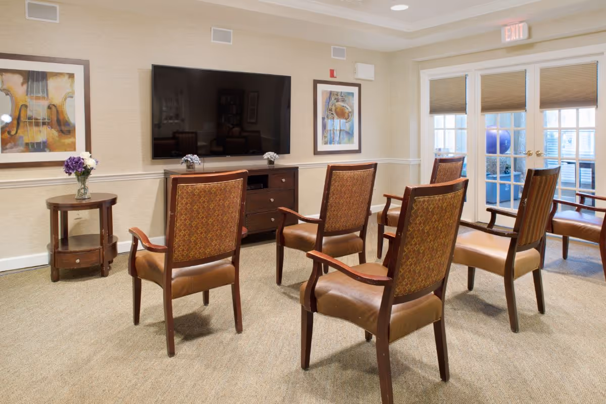 A cozy common area with six wooden armchairs arranged in two rows facing a large flat-screen TV mounted on the wall. The room has beige walls with two framed paintings on either side of the TV, a small round wooden side table with a vase of flowers, and double glass doors with blinds leading to another room.