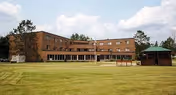 Front view of a three-story brick senior living building with a large grassy lawn and a gazebo under a partly cloudy sky.
