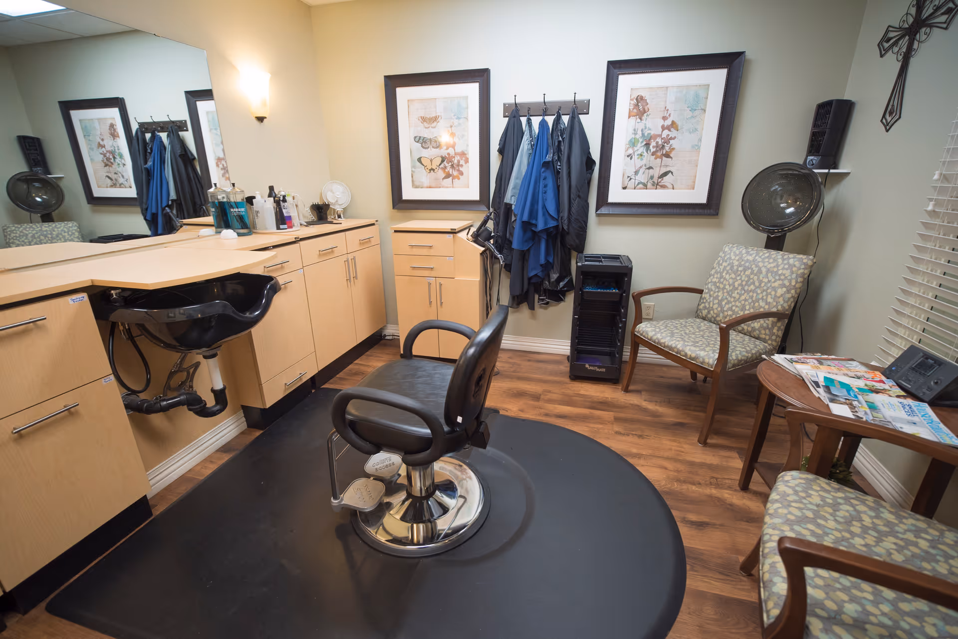 Interior of a hair salon room with a black salon chair on a black mat, a black hair washing sink, wooden cabinets with hair products on the countertop, a coat rack with several capes hanging, two framed floral and butterfly pictures on the wall, a patterned armchair, a small table with magazines, and a hair dryer hood in the corner.