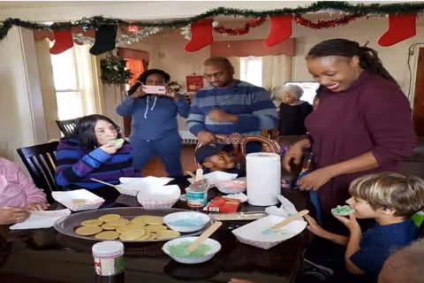 A group of people, including children and adults, gathered around a table in a festive room decorated with Christmas stockings and garlands. They are engaged in decorating cookies with green icing and enjoying the activity together.