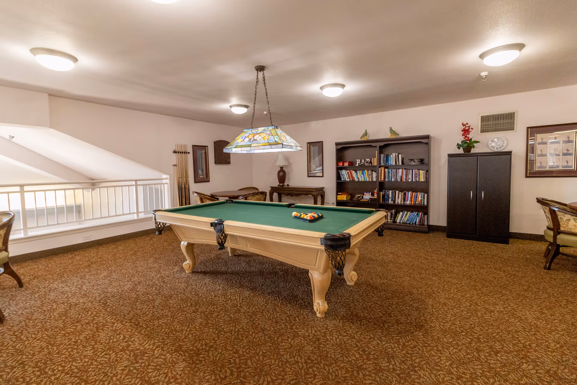 A recreational room with a pool table in the center, a hanging stained glass light fixture above it, bookshelves filled with books against the wall, and several chairs and tables around the room. The room has carpeted flooring and neutral-colored walls.