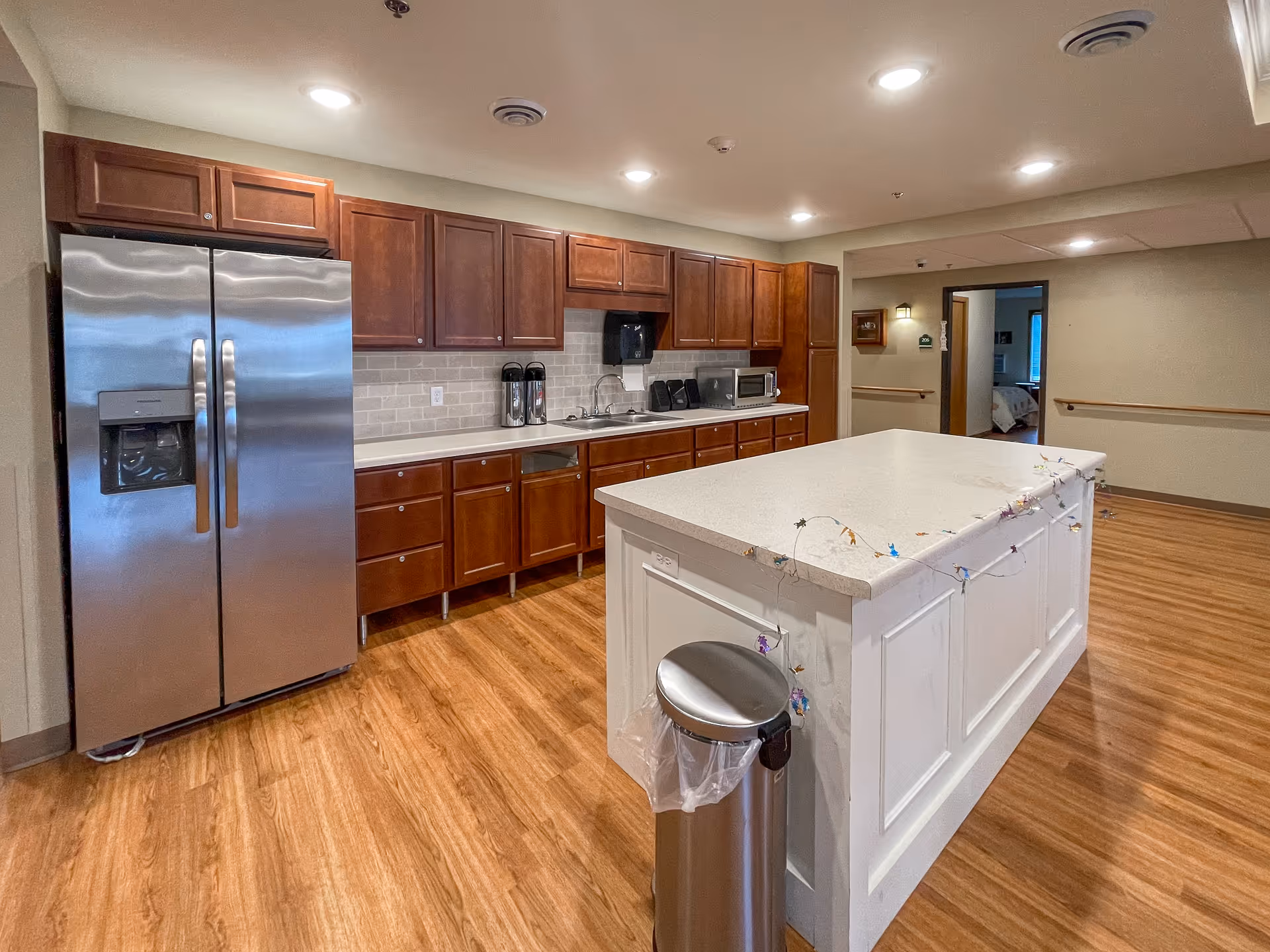 A spacious communal kitchen with a large white island, stainless steel refrigerator, wooden cabinets, and countertop appliances.