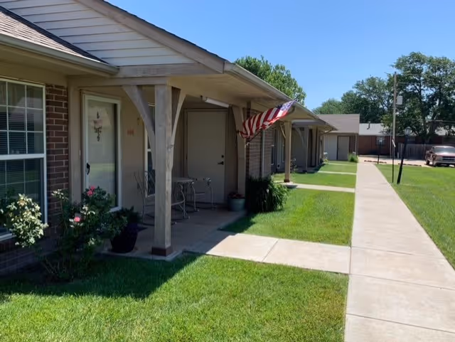 Exterior view of a single-story residential building with a covered porch, an American flag hanging near the entrance, green grass lawn, and a concrete walkway leading past the units under a clear blue sky.