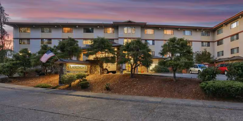 Exterior view of a multi-story senior living facility building at dusk with lights on inside. The building is surrounded by trees and landscaping, with a sign in front that reads 'CLAREMONT'. Several cars are parked near the entrance.