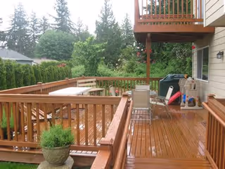 Wooden outdoor deck attached to a house with patio furniture including a table, chairs, a grill, and a small playhouse. The deck is surrounded by a wooden railing and overlooks a green garden area with trees and shrubs.
