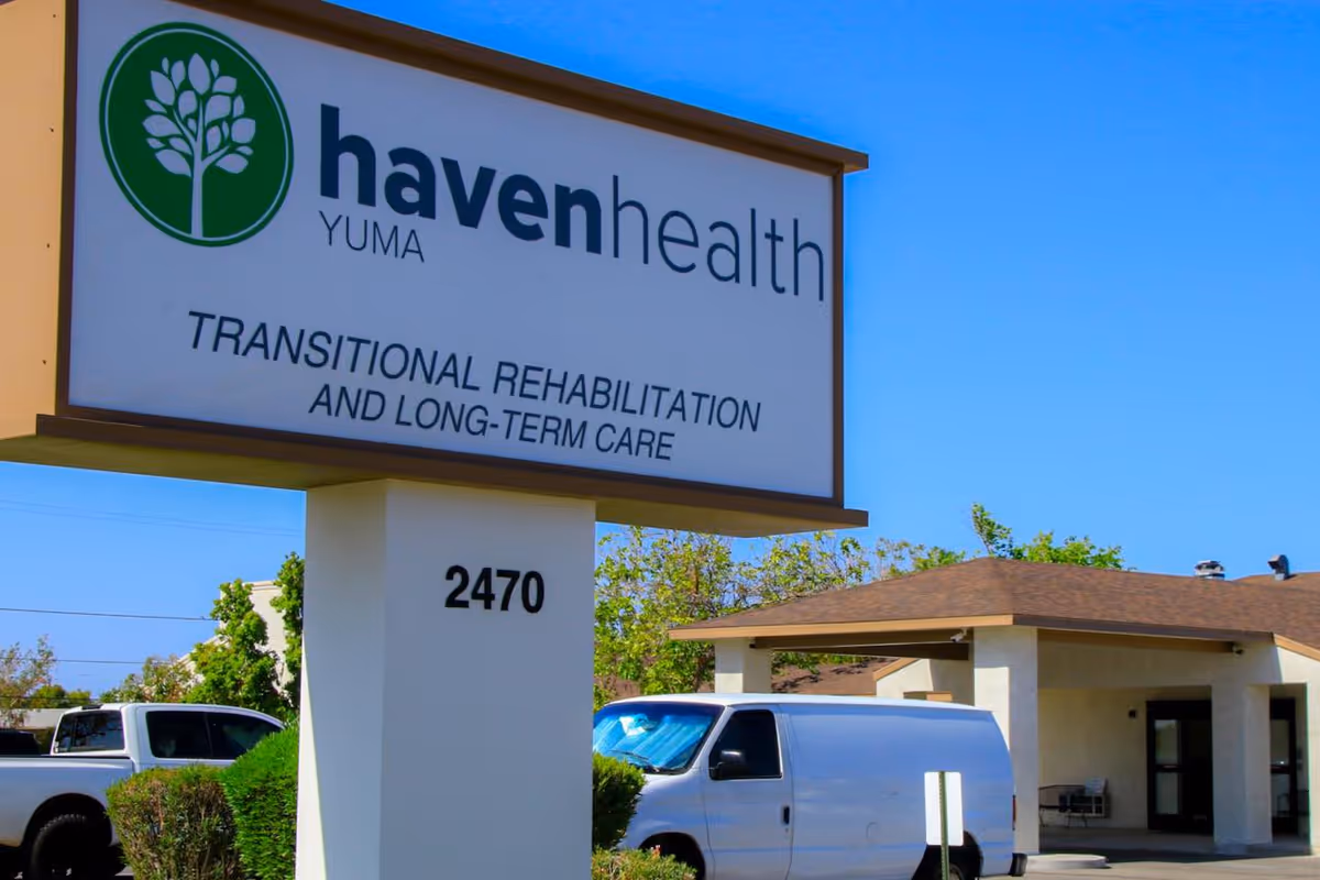Outdoor view of the Haven Health Yuma facility sign displaying 'Transitional Rehabilitation and Long-Term Care' with the address number 2470. In the background, there is a building with a brown roof, some greenery, and parked vehicles including a white van and a white truck under a clear blue sky.