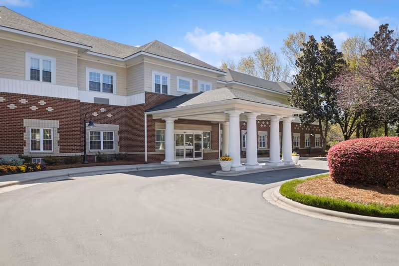 Exterior view of a senior living facility building with a covered entrance supported by white columns, surrounded by landscaped bushes and trees under a clear blue sky.