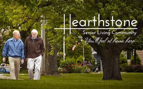 Two older men walk across a grassy, tree-lined outdoor area with landscaping and an overlaid "Hearthstone Senior Living Community — You'll feel at home here" logo.