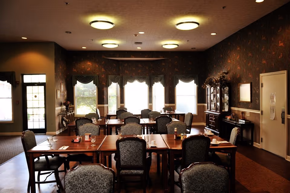 Dining room with multiple set wooden tables and upholstered chairs, large windows with valances, and a buffet cabinet along the right wall.