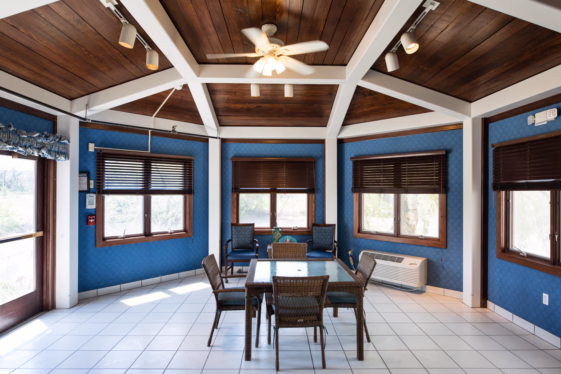 Sunlit common room with a glass-top dining table and wicker chairs beneath a wood-beamed ceiling and multiple windows.