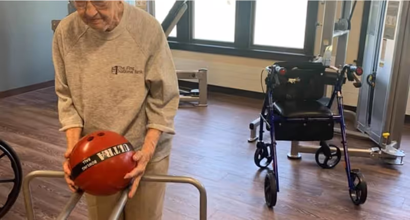 An elderly person wearing a light gray sweatshirt is holding a red bowling ball in a room with wooden flooring. There is a walker with a black seat and a piece of exercise equipment visible in the background near a window.