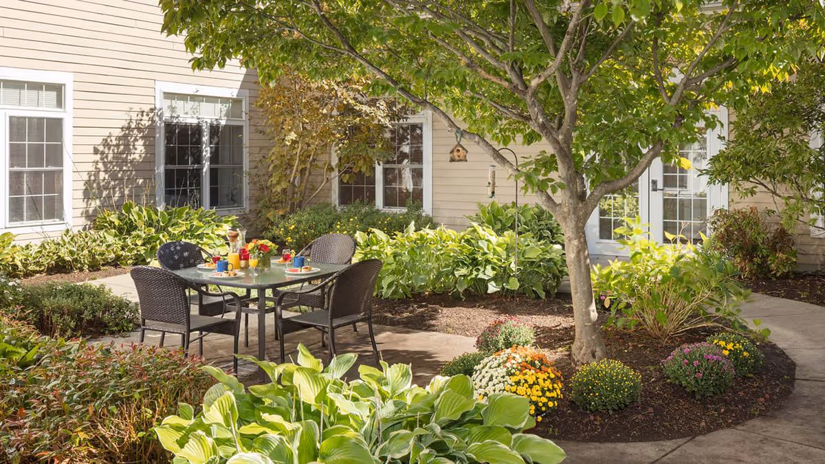 Outdoor patio area at Atria Bay Spring Village featuring a glass-top table with four black wicker chairs. The table is set with colorful cups, plates, and a pitcher of orange juice. Surrounding the patio are lush green plants, bushes, and a tree providing shade. The building exterior with beige siding and white-framed windows and doors is visible in the background.