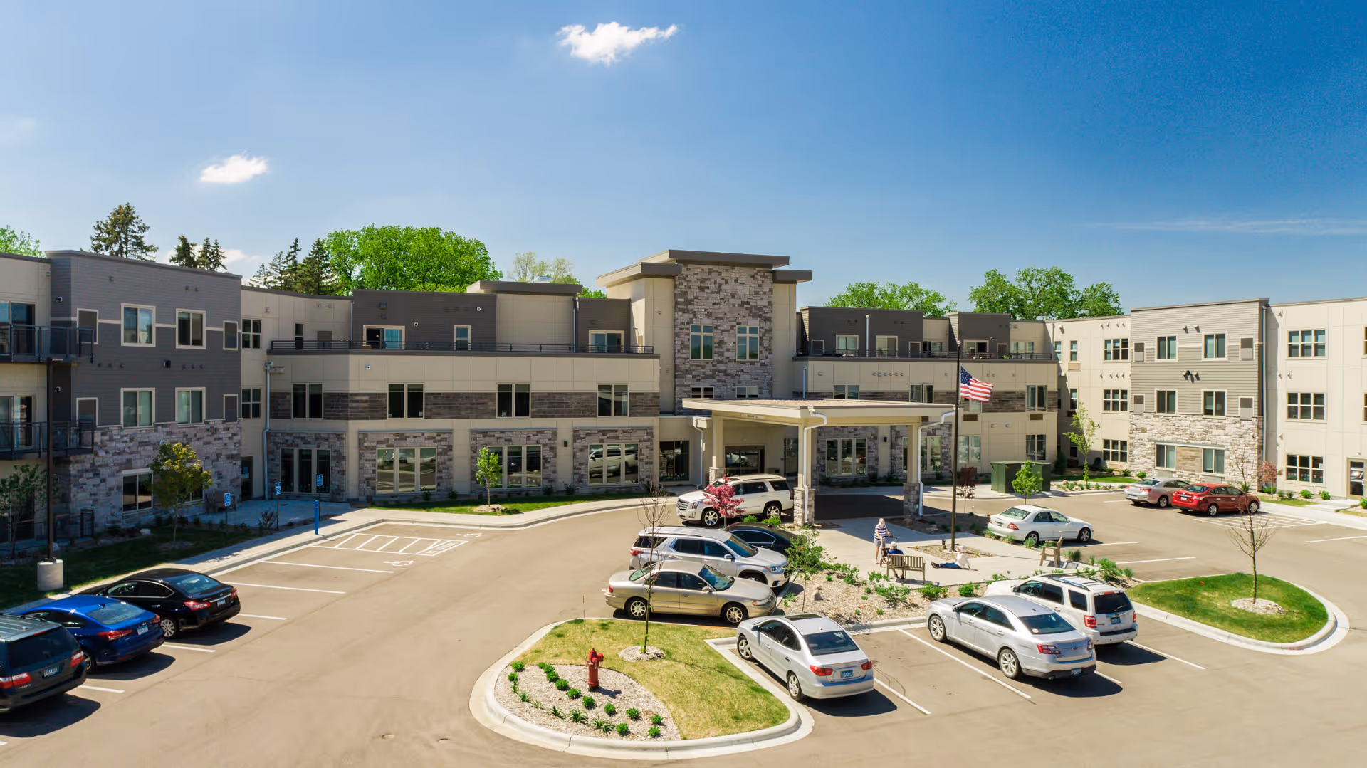Exterior view of Havenwood of Minnetonka, a modern three-story senior living facility with a covered entrance, multiple windows, and a parking lot with several cars parked. The sky is clear and blue with a few clouds, and there are trees and landscaping around the building.