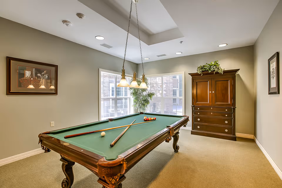 A well-lit game room with a green felt pool table at the center, two pool cues resting on the table, and a rack of billiard balls arranged near one end. The room has beige walls, a large window letting in natural light, a wooden cabinet with drawers and a plant on top, and framed artwork on the walls. A hanging light fixture with three lamps is positioned above the pool table.
