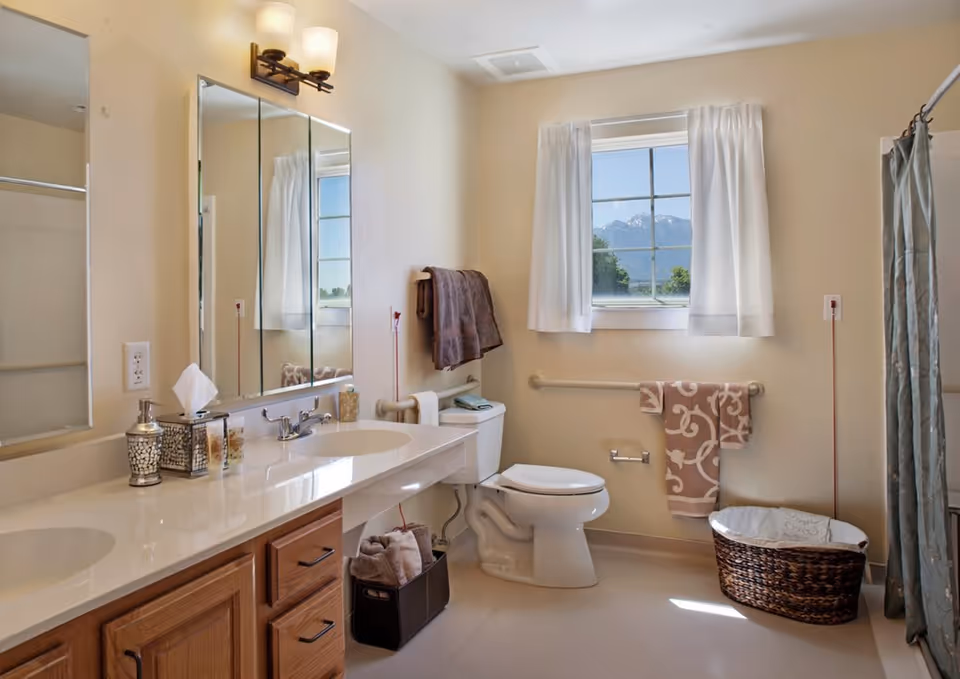A clean and well-lit bathroom with a double sink vanity, large mirror, toilet, and a window with white curtains showing a mountain view. There are towels hanging on the wall, a wicker laundry basket, and a shower with a curtain on the right side.
