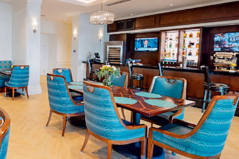 Bright dining area with teal upholstered chairs around wooden tables and a bar with TVs in the background.