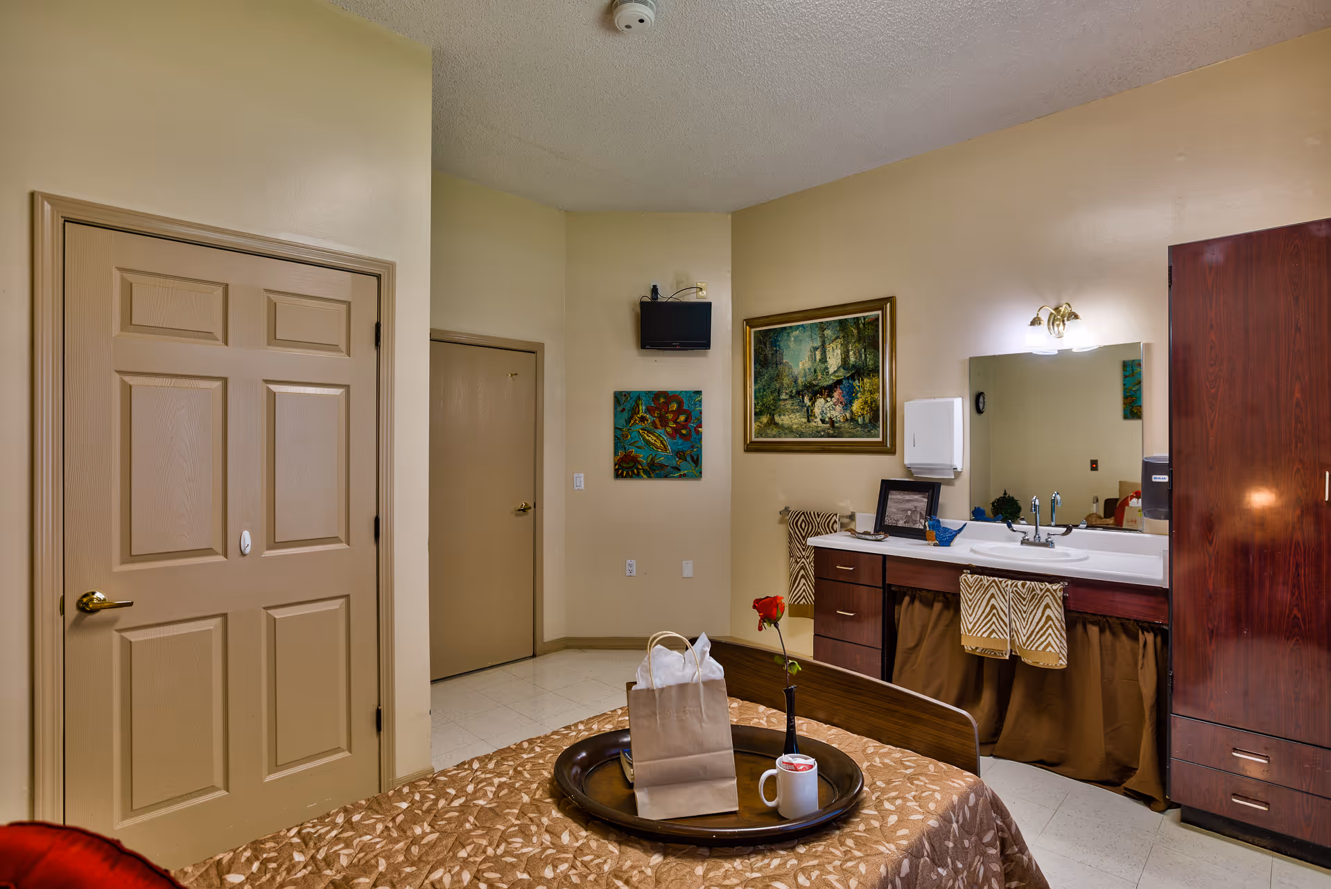 Interior view of a senior living facility room at The Hilltop at Blue River, featuring a bed with a brown patterned bedspread and a tray holding a paper bag, a coffee mug, and a single red rose in a vase. The room includes a sink with a mirror above it, a towel rack with patterned towels, a wooden cabinet, and two paintings on the wall. There are two closed doors visible, and the walls are painted beige.