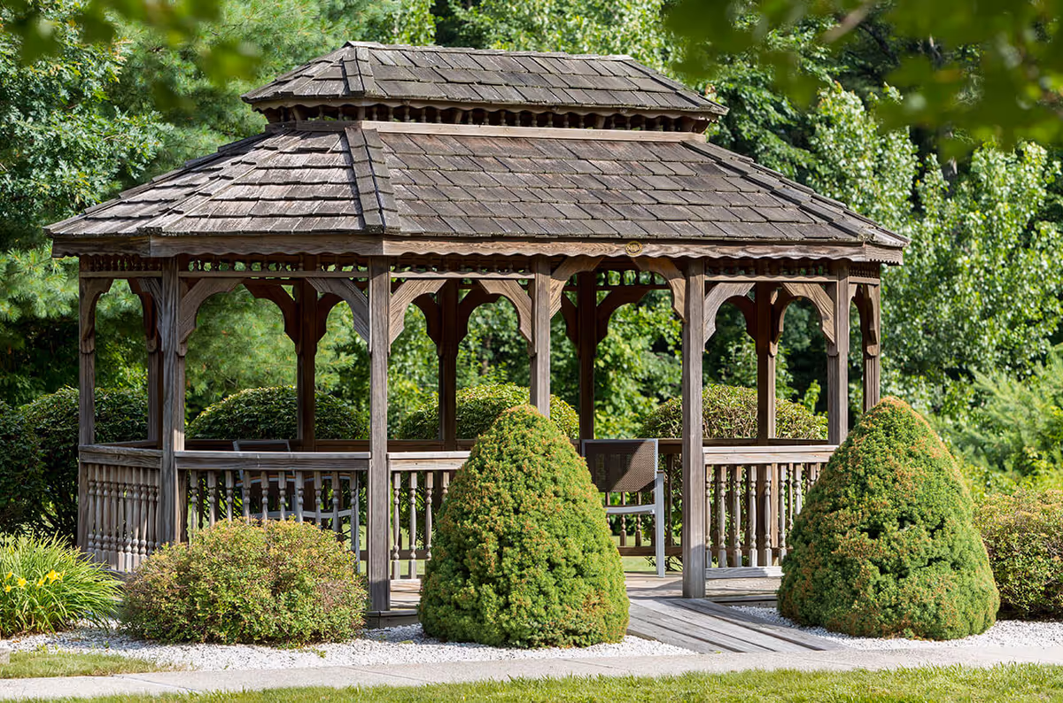 A wooden gazebo with a shingled roof surrounded by neatly trimmed bushes and greenery in a garden setting at Benchmark at Orchard Valley.