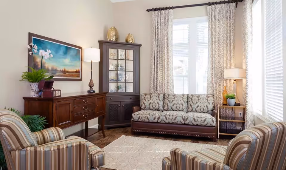 Bright furnished living room with a patterned sofa, two striped armchairs, wooden sideboard and cabinet, and large windows with curtains.