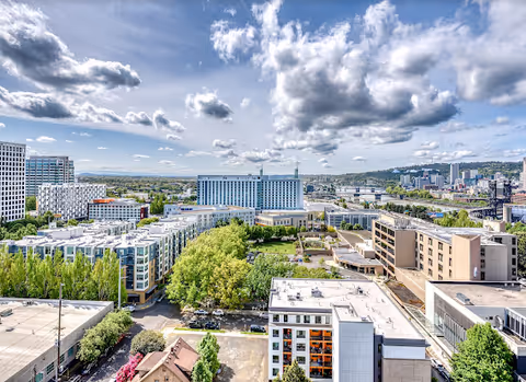 A high-angle view of an urban area with multiple mid-rise buildings, green trees, and a partly cloudy sky. The scene includes residential and commercial buildings with a river and distant hills in the background.