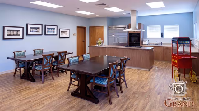 A spacious dining area with two dark wooden tables surrounded by chairs with blue striped cushions. The room features light blue walls adorned with framed pictures, a kitchen area with wooden cabinets, stainless steel appliances, and a popcorn machine on the right side. The floor is wooden, and the room is well-lit with ceiling lights and natural light from two windows.