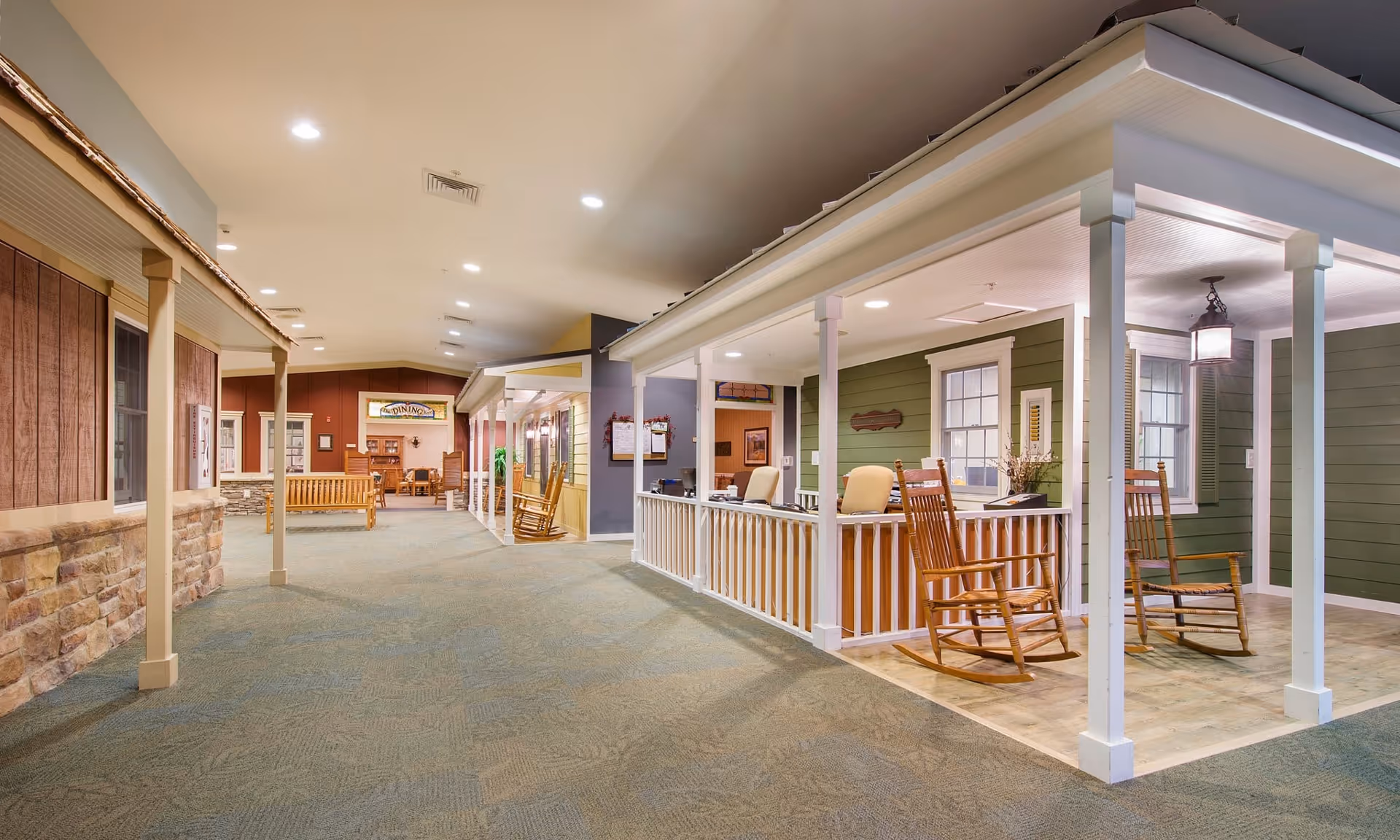 Indoor hallway designed to look like an outdoor street with house facades, including porches with rocking chairs and benches, under bright ceiling lights.