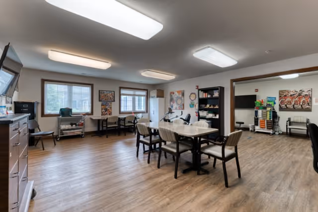 A spacious room with wooden flooring and multiple tables and chairs arranged for group activities or meetings. The room has large windows allowing natural light, fluorescent ceiling lights, and various decorations on the walls. There is a bookshelf with supplies and another adjoining room visible through a wide doorway, which contains additional chairs and storage carts.