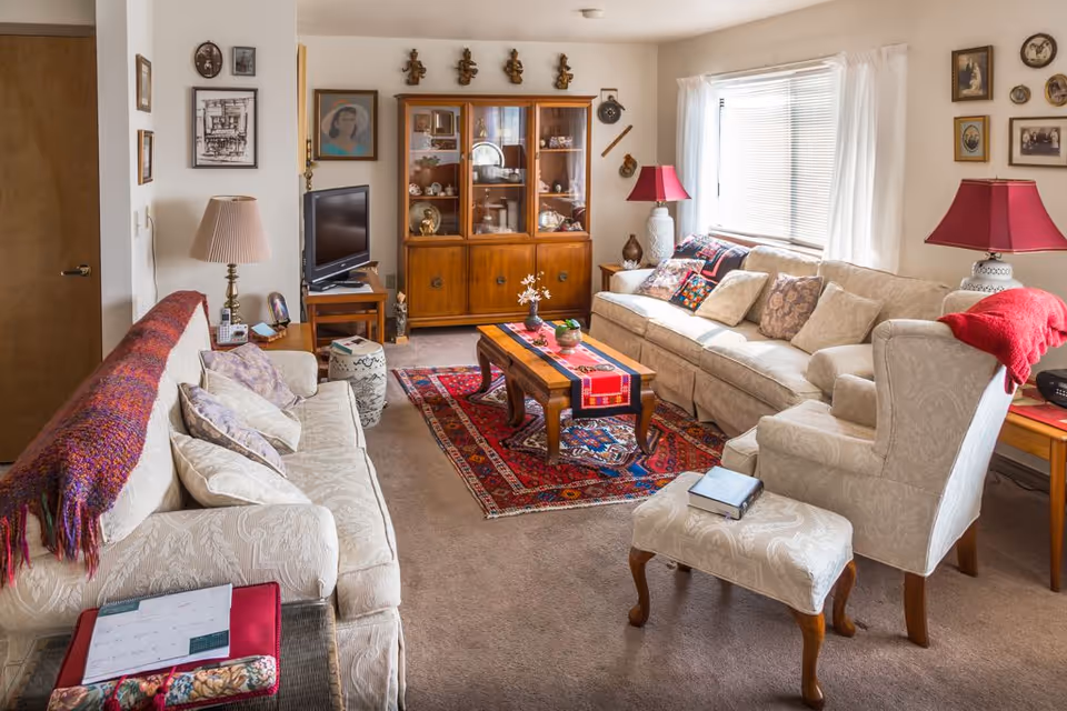A cozy living room with two beige sofas, an armchair with an ottoman, a wooden coffee table on a colorful rug, and a wooden cabinet with glass doors displaying decorative items. The room is decorated with framed pictures on the walls, two table lamps with red shades, and a window with white curtains letting in natural light.